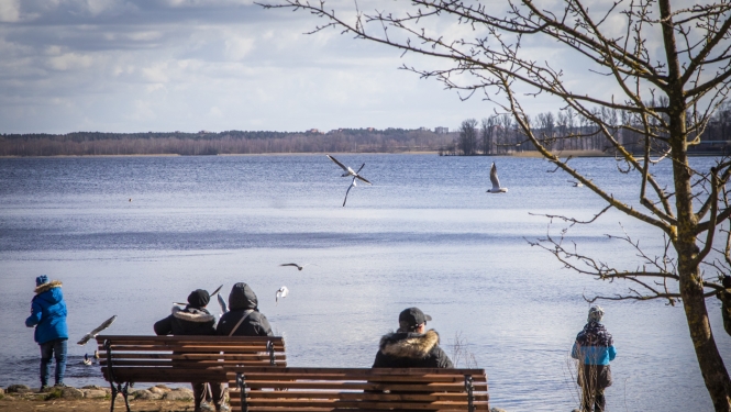Cilvēki sēž uz soliņiem ezera krastā