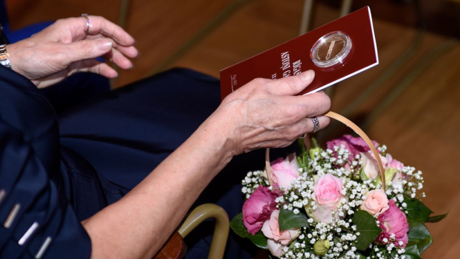 Rokās tur ziedus un jubilejas sudraba monētu